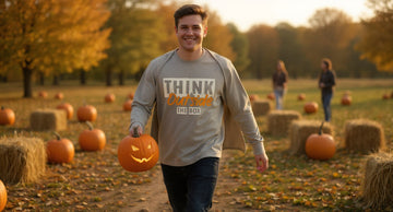 Person walking through a pumpkin patch at golden hour wearing a sand typography tee, carrying a carved pumpkin, a friend blurred behind them.