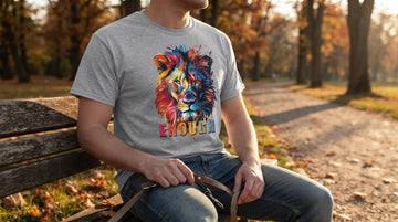 A person in a heather grey lion tee sits on a park bench at golden hour, hand resting on a dog leash amid falling leaves.