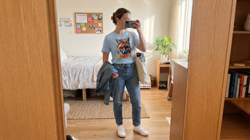 Person in a light blue fox graphic tee taking a morning mirror selfie in a bright bedroom with jeans, white sneakers, and a canvas tote.