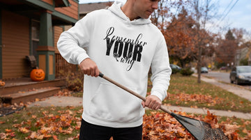 A person in a white hoodie rakes orange leaves beside a porch with a carved pumpkin.