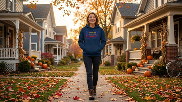 A young woman smiles while walking down a sidewalk lined with fall leaves and porch pumpkins, wearing a navy blue hoodie.
