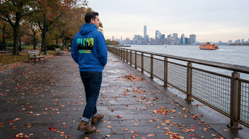 Person in a Royal blue hoodie walking beside fallen leaves on a Staten Island waterfront promenade with ferry and skyline beyond.