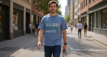 A person in a graphic tee and straight jeans walks down a sunlit city street carrying a tote bag, storefronts behind.