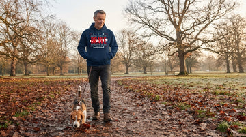 A man in his late fifties walks a beagle on a leaf-covered park path while wearing a dark hoodie.