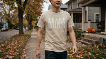 A person in a graphic tee walks along a tree-lined street with fallen leaves and pumpkins on porch steps.