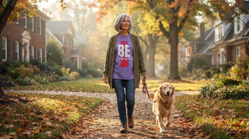 Gray-haired woman in a cardigan walks a dog on a leaf-covered neighborhood path while wearing a visible graphic tee.