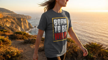 A person wears the Born To Be Wild T-shirt on a coastal trail with ocean cliffs behind them at golden hour.