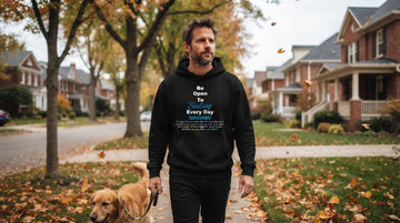 A man in a black printed hoodie walks a dog on a leaf-covered suburban street during an overcast autumn morning.