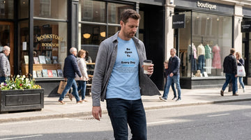 Man in late thirties walking on a city street wearing a light blue t-shirt under an open grey overshirt.