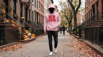 Person in a light pink hoodie walks along a brick-lined sidewalk with fallen leaves and pumpkins on stoops.