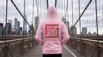 Person wearing a light pink Bronx hoodie walking across a Brooklyn Bridge walkway on a crisp November morning.