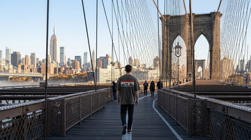 A person wearing a grey Brooklyn 1816 t-shirt walks along the Brooklyn Bridge promenade with the city skyline behind them.
