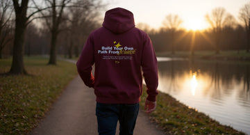 Person in a maroon hoodie walking a quiet lakeside path on a cool autumn morning, hands in the front pocket, bare trees behind.