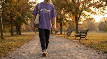Person in a violet printed T-shirt walks on a gravel park path with a tote bag during golden autumn light.
