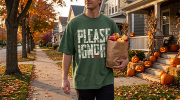 A person in a printed t-shirt carries a paper bag of apples down a sunny neighborhood sidewalk lined with carved pumpkins.